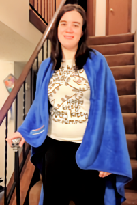 Christina standing on the stairs holding her skull-tipped cane with a fuzzy blue blanket draped over her shoulders like a cloak